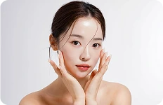 Młoda kobieta o gładkiej cerze pozująca z dłońmi przy twarzy na jasnym tle.