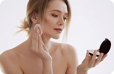 Kobieta nakładająca podkład typu cushion za pomocą różowej gąbeczki, patrząca w lusterko kompaktowe.