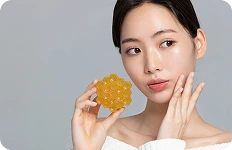 Młoda kobieta o promiennej cerze trzymająca przy twarzy żółty przedmiot w kształcie plastra miodu na szarym tle.