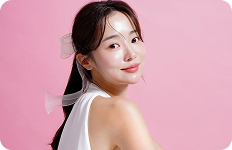 Młoda kobieta o jasnej, promiennej cerze i ciemnych włosach spiętych w kucyk z dużą, półprzezroczystą kokardą, pozująca na jasnoróżowym tle.