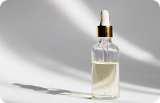Przezroczysta szklana buteleczka z zakraplaczem, wypełniona jasnożółtym olejkiem lub serum, stojąca na jasnym tle w naturalnym świetle.