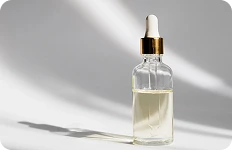 Przezroczysta szklana buteleczka z zakraplaczem, wypełniona jasnożółtym olejkiem lub serum, stojąca na jasnym tle w naturalnym świetle.