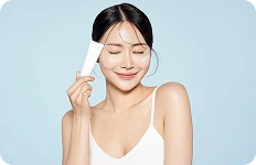 Uśmiechnięta kobieta trzymająca białą tubkę kremu, z odrobiną kosmetyku nałożoną na policzek, na jasnoniebieskim tle.